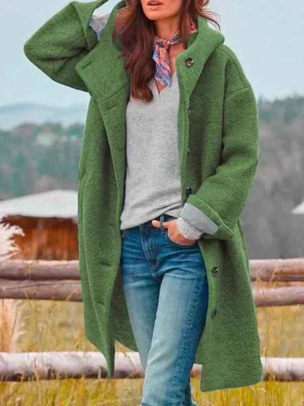 Mantel in Grün mit Kapuze und Knöpfen, getragen zu grauem Pullover, Halstuch und Jeans, Wiese und Holzzaun im Hintergrund. 5