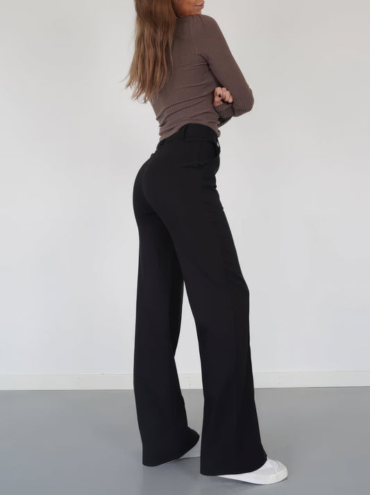 Frau in schwarzer Schlaghose und braunem Oberteil, seitlich, weiße Sneaker, minimalistischer Stil.
