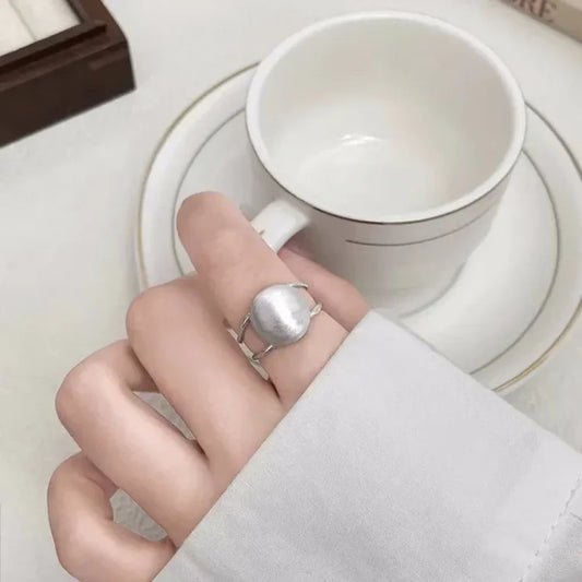 Silberner Ring mit großer Perle auf Hand, weiße Tasse im Hintergrund, eleganter Schmuck.
