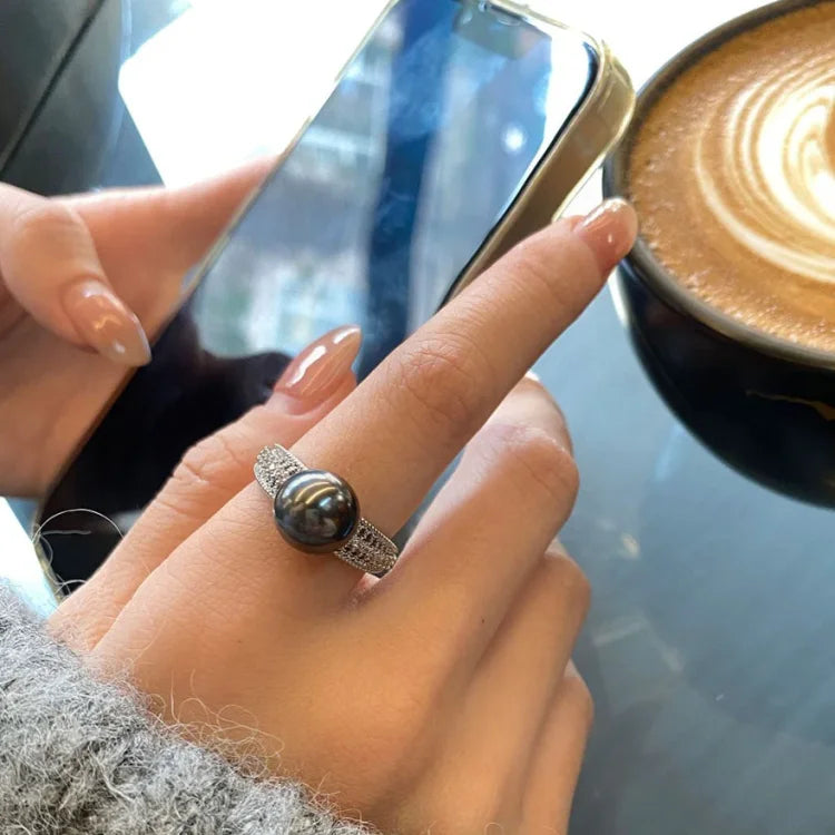 Frau mit schwarzem Perlenring, Smartphone und Cappuccino, eleganter Schmuck, Nahaufnahme.