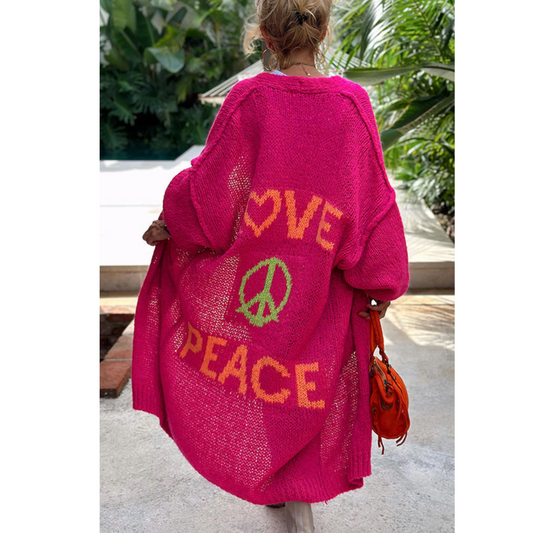 Frau in pinkem Strickmantel mit "Love Peace" Schriftzug, tropische Umgebung.