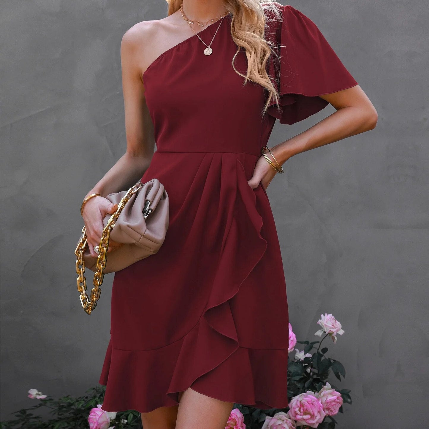 Frau in burgunderrotem One-Shoulder-Kleid mit Tasche, elegante Mode, Blumenhintergrund.