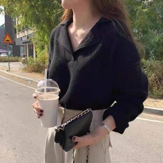 Frau in schwarzem Pullover mit Kette, hält Getränk und schwarze Tasche, draußen auf Straße.