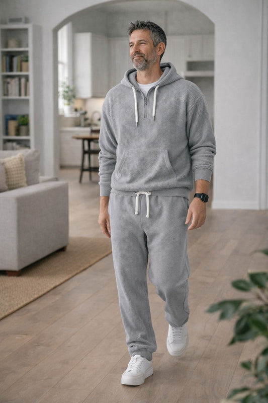Mann in hellgrauem Waffelstrick-Set mit Hoodie und Jogginghose läuft durch ein helles Wohnzimmer. 2