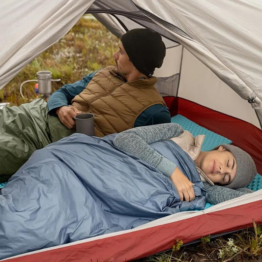 Zwei Camper im Zelt, schlafend in blauen Schlafsäcken, mit Mützen und Thermobechern.