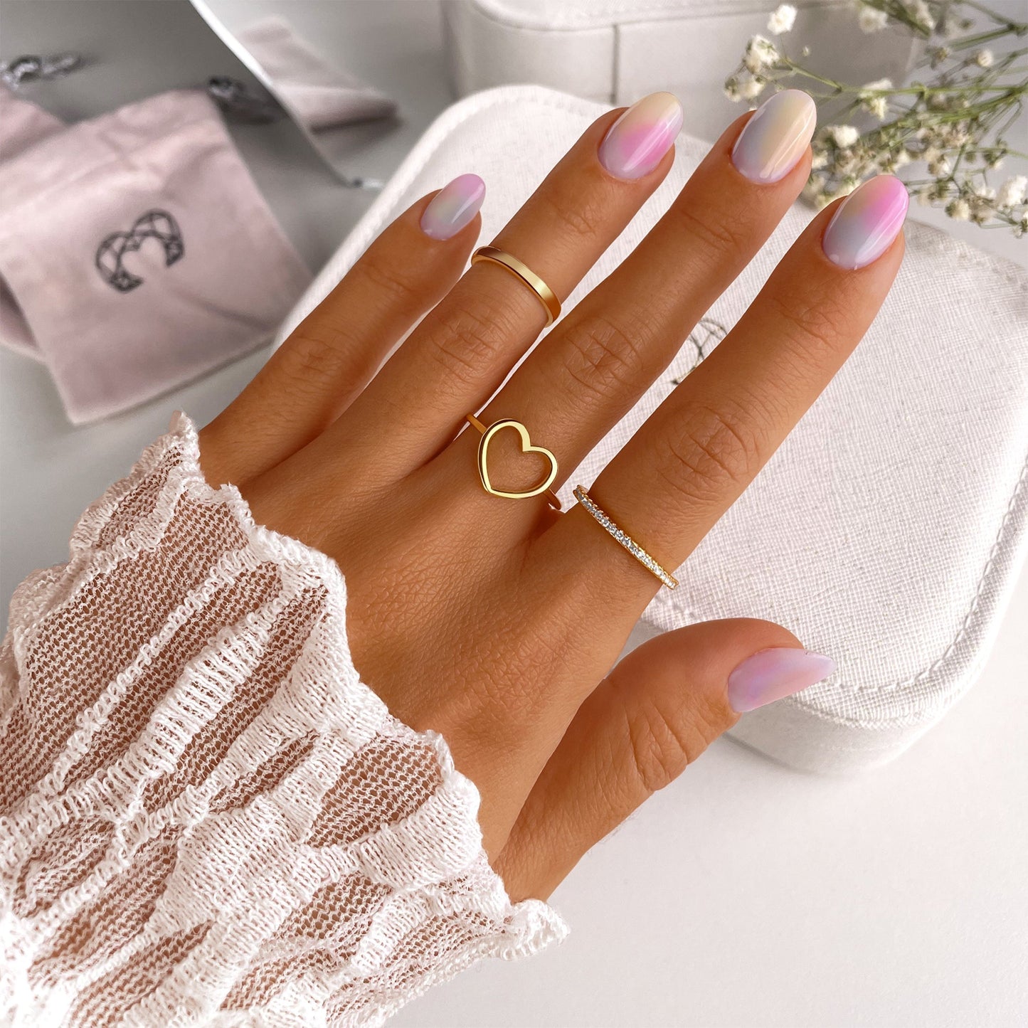 Damenhand mit Herzring, Goldring und Diamantring, pastellfarbene Nägel, eleganter Schmuck.