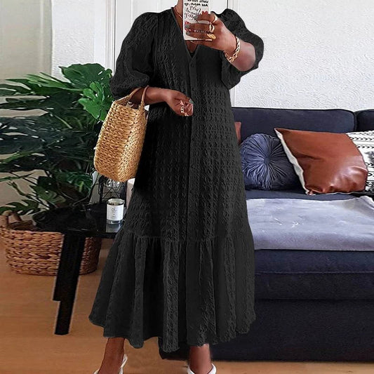Frau in schwarzem Maxikleid mit Strohtasche vor Sofa und Pflanzen im Wohnzimmer.