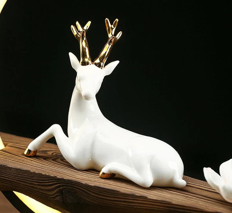 Weiße Keramikfigur eines liegenden Hirsches mit goldenen Geweihspitzen auf Holzbrett.