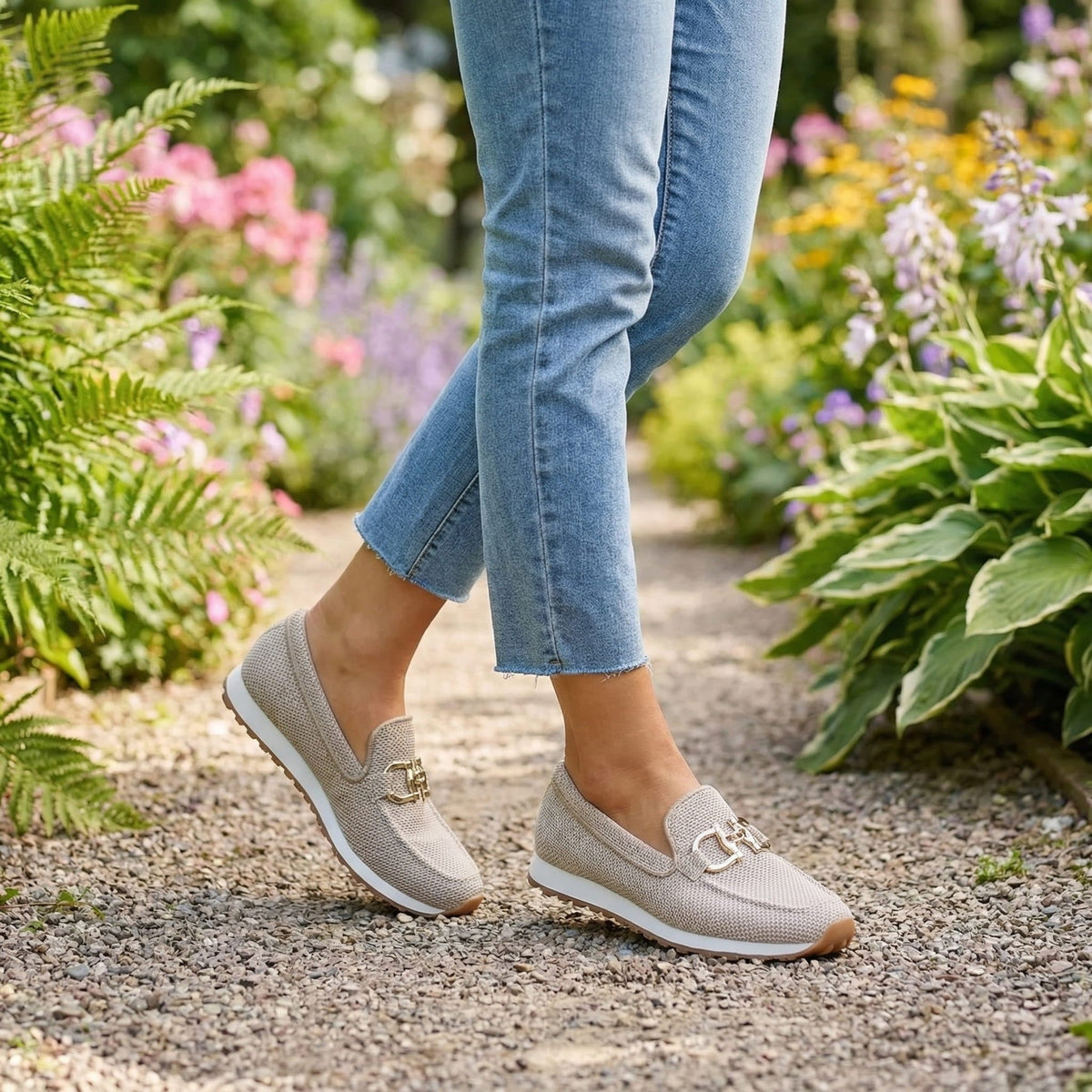Beigefarbene Slipper mit Jeans beim Gehen auf einem Kiesweg im blühenden Garten 11