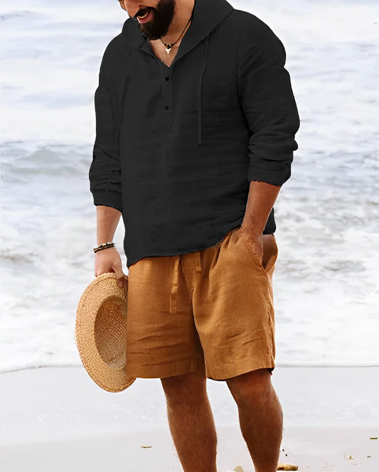 Mann in schwarzem Leinenhemd und braunen Shorts am Strand, hält Strohhut.