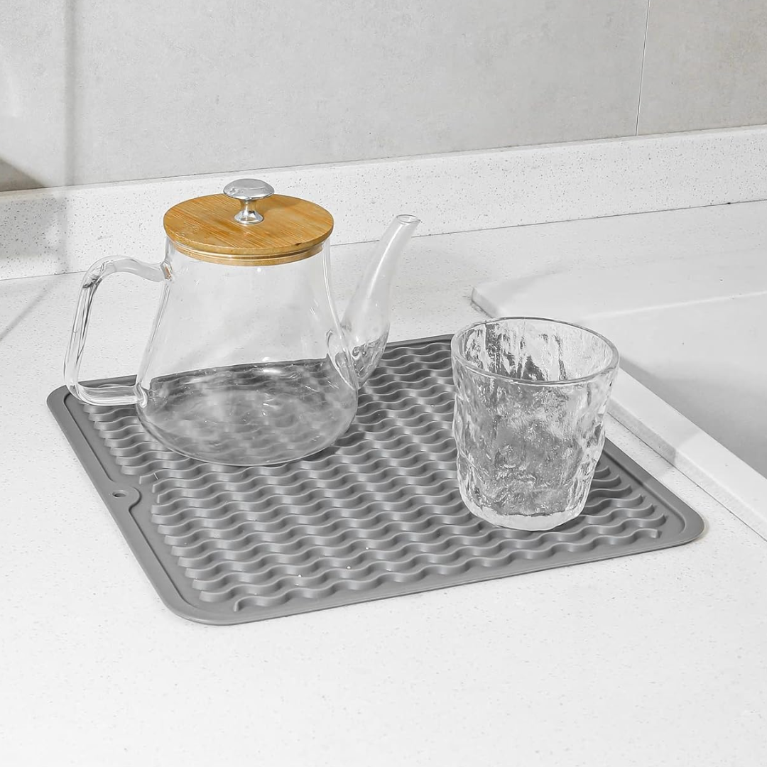 Glas-Teekanne mit Holzdeckel und Glas auf grauer Silikon-Abtropfmatte in moderner Küche.