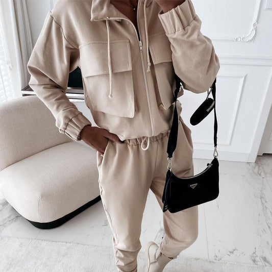 Beige Damen-Trainingsanzug mit Kapuze, Reißverschluss und schwarzer Umhängetasche im Wohnzimmer.