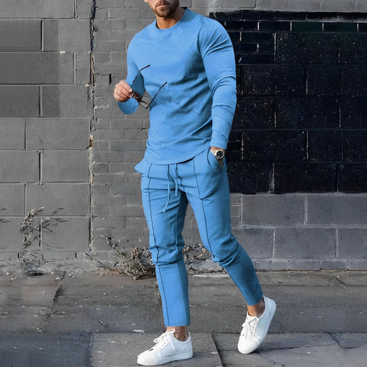 Mann in blauem Jogginganzug und weißen Sneakers vor grauer Wand, hält Sonnenbrille.