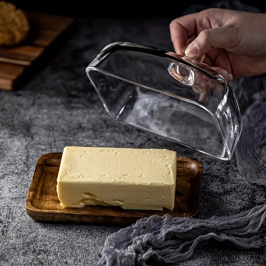 Hand hebt Glas-Butterdose, Holzbrett, Butterstück, Küche, Lebensmittelaufbewahrung.