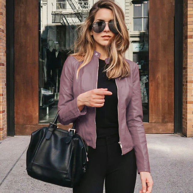 Frau in lila PU-Lederjacke, schwarze Hose, Sonnenbrille, schwarze Tasche, urbaner Hintergrund.