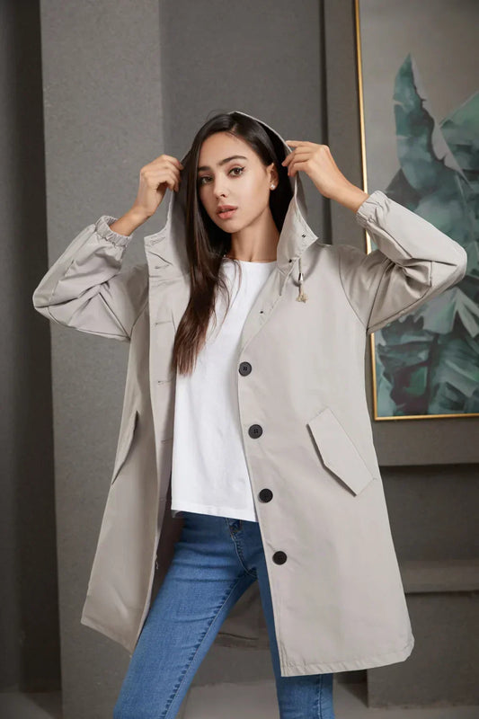 Frau in beigem Regenmantel mit Kapuze, weiße Bluse, blaue Jeans, moderne Mode.