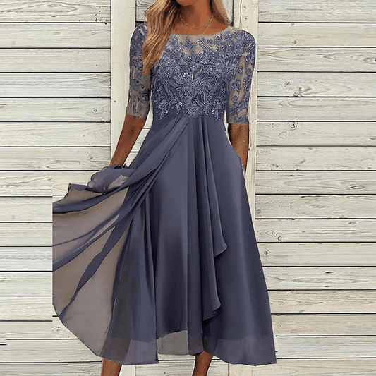 Frau in elegantem, blauem Sommerkleid mit Spitze, knielang, vor Holzwand.