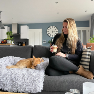 Frau mit Hund auf grauem Sofa, hält Kaffee, modernes Wohnzimmer, gemütliche Atmosphäre.