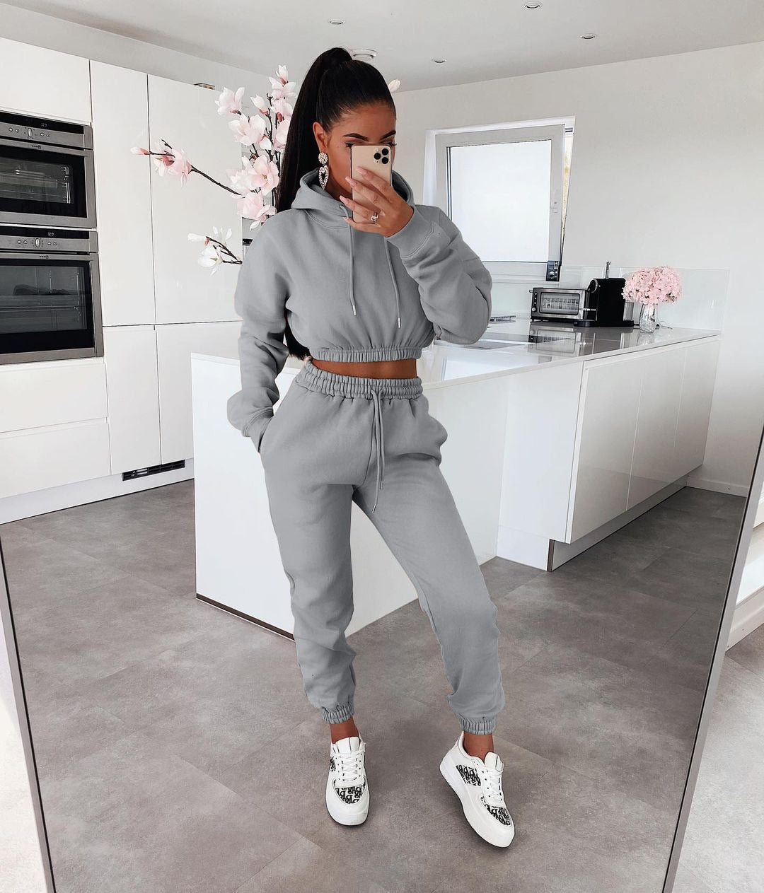 Frau in grauem Jogginganzug in moderner Küche, trägt weiße Sneaker, macht Selfie.