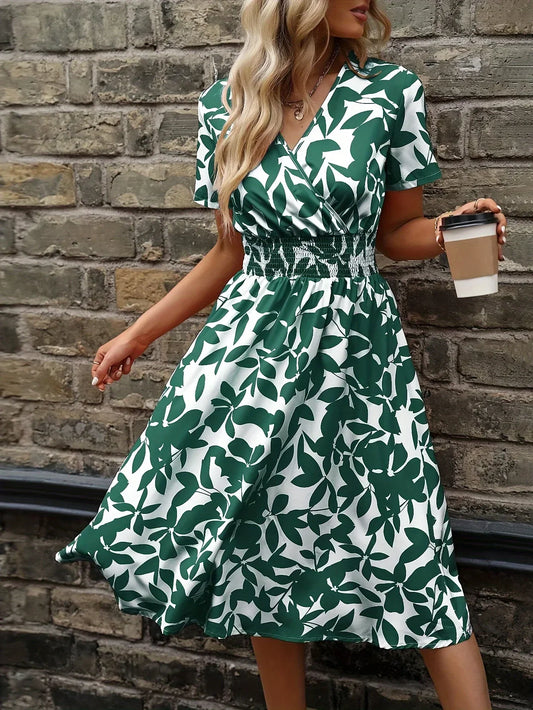 Frau im grünen Sommerkleid mit Blattmuster hält Kaffeebecher vor Backsteinmauer.