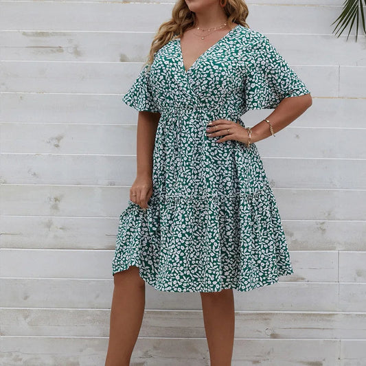 Frau in grünem Sommerkleid mit Blumenmuster vor weißer Holzwand.