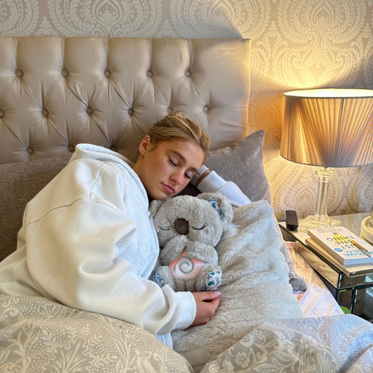 Frau schläft im Bett mit Plüsch-Koala, beige Bettwäsche, Nachttischlampe, gemütliches Schlafzimmer.