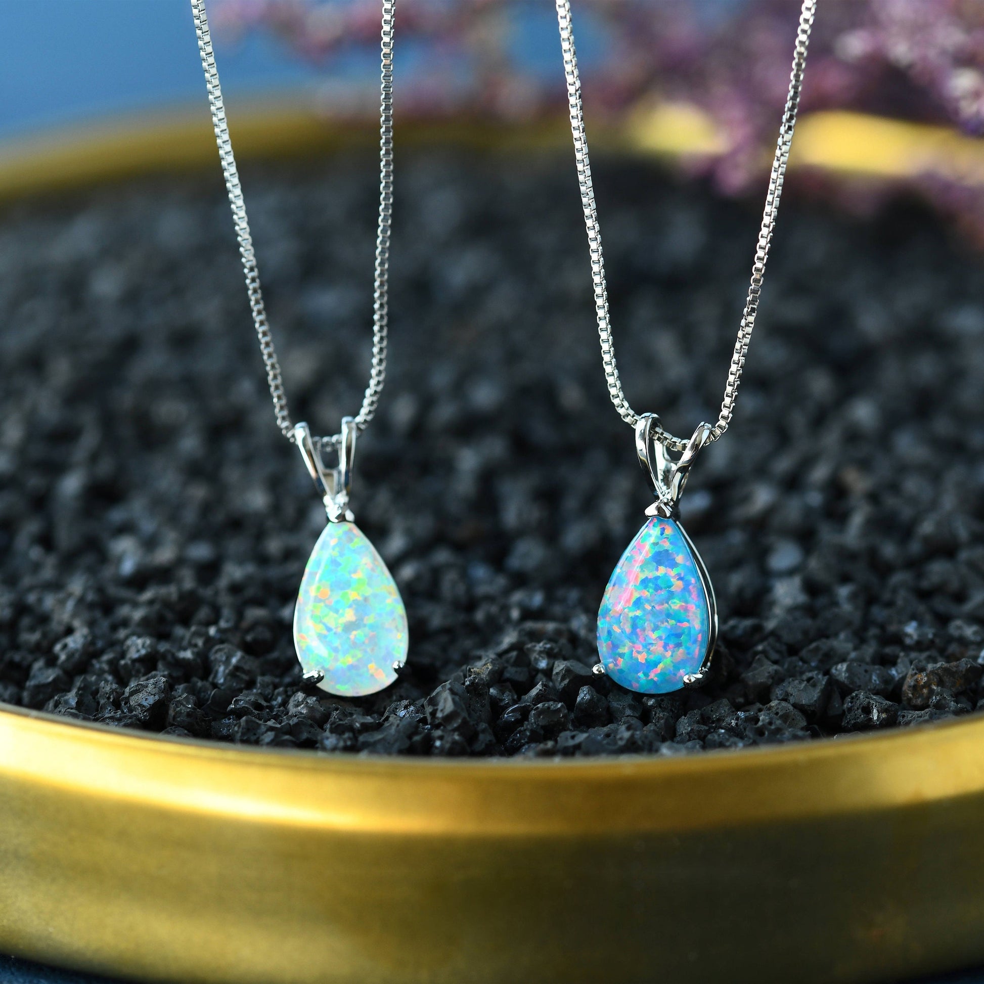 Zwei Opal-Anhänger an Silberketten auf schwarzem Kies in goldener Schale, eleganter Schmuck.
