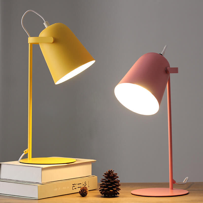 Zwei eingeschaltete Tischlampen im Vergleich, eine gelbe auf Büchern und eine rosafarbene auf dem Holztisch, daneben ein Tannenzapfen. 5