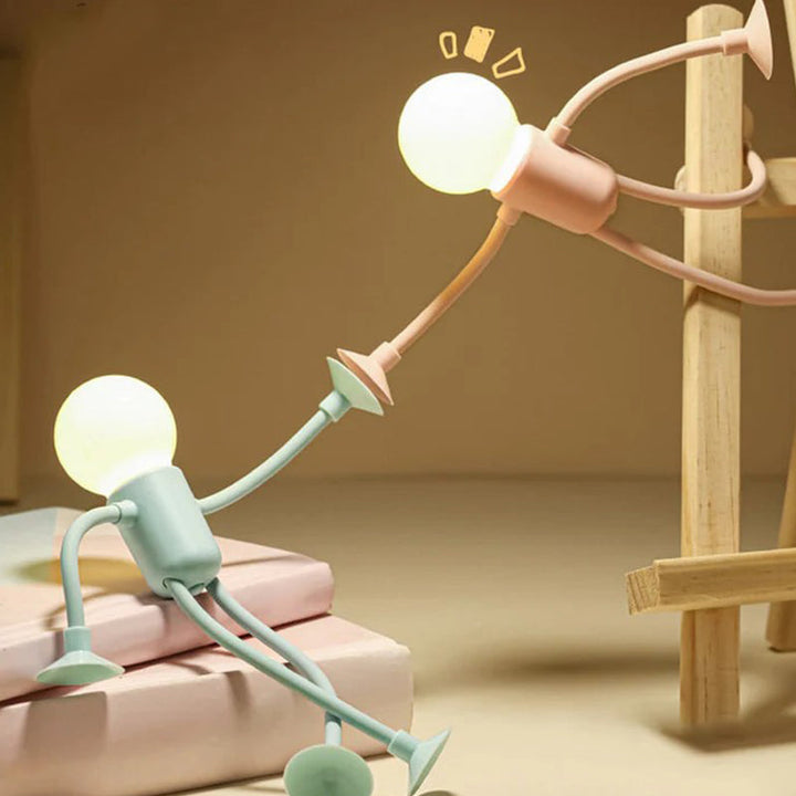 Grünes Männchen-Licht sitzt auf zwei Büchern und streckt den Arm einem rosa Licht entgegen, das an einem Holzgestell hängt. 6