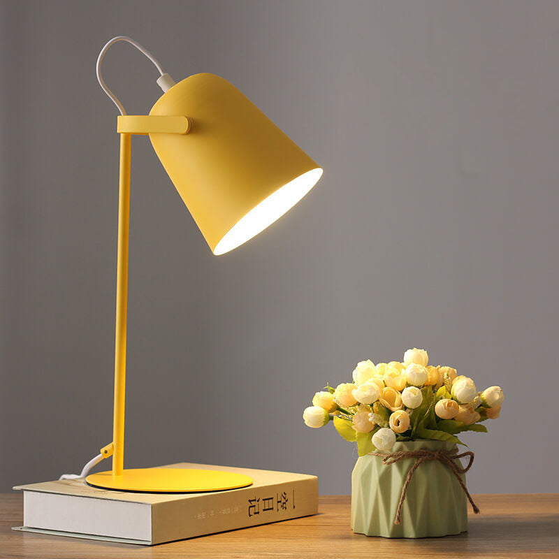 Gelbe Tischlampe mit eimerförmigem Schirm leuchtet auf einem Buch, daneben eine kleine Vase mit hellen Röschen auf einem Holztisch. 1