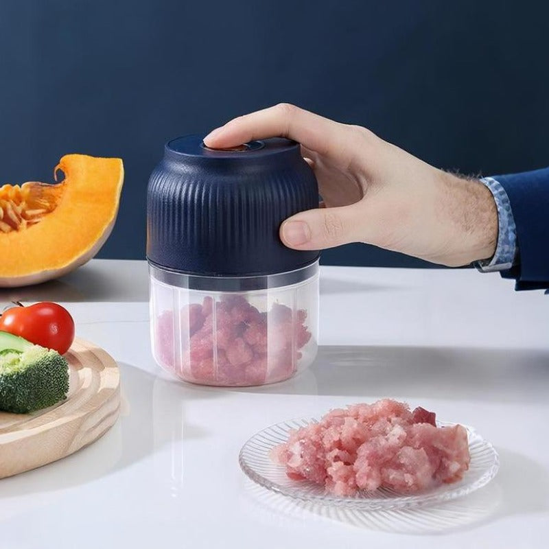 Eine Hand drückt auf den dunkelblauen Deckel des Mini-Hackers, in der durchsichtigen Schüssel liegt gehacktes Hackfleisch, daneben Kürbis, Tomate und Brokkoli. 1