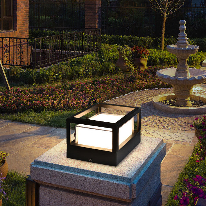 Eckige schwarze Leuchte mit offenem Rahmen auf einem Granitpfeiler in einem abendlichen Garten mit Springbrunnen und Blumenbeeten. 2