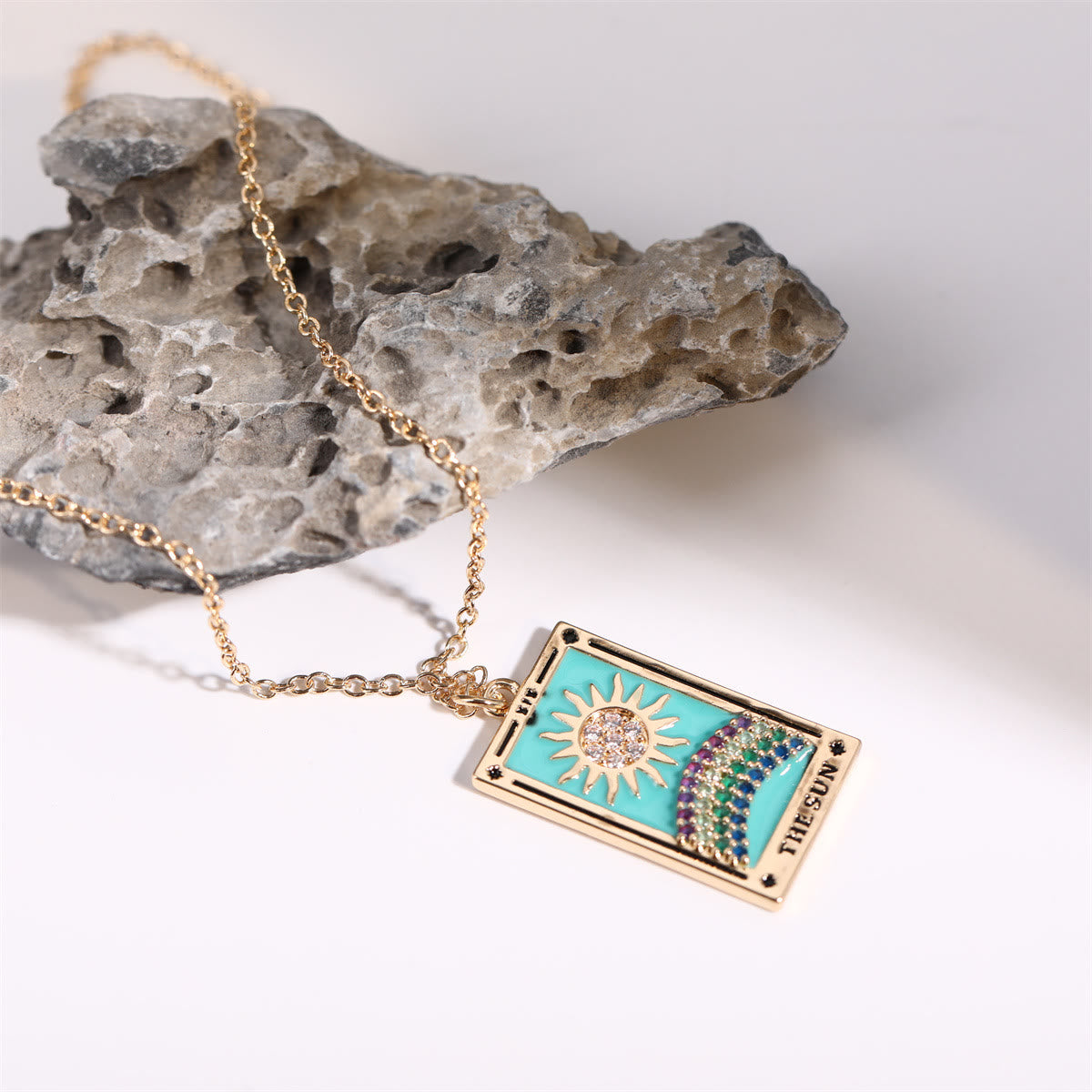 Goldene Halskette mit Tarot-Anhänger, Sonnenmotiv, türkisfarben, auf grauem Stein.