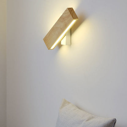 Schräg gedrehte Holz-Wandlampe mit leuchtender LED-Leiste an einer hellen Wand über einem beigen Kissen. 1