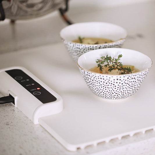 Zwei Schalen mit Suppe auf weißem Tisch, dekoriert mit Thymian, neben digitalem Heizgerät.