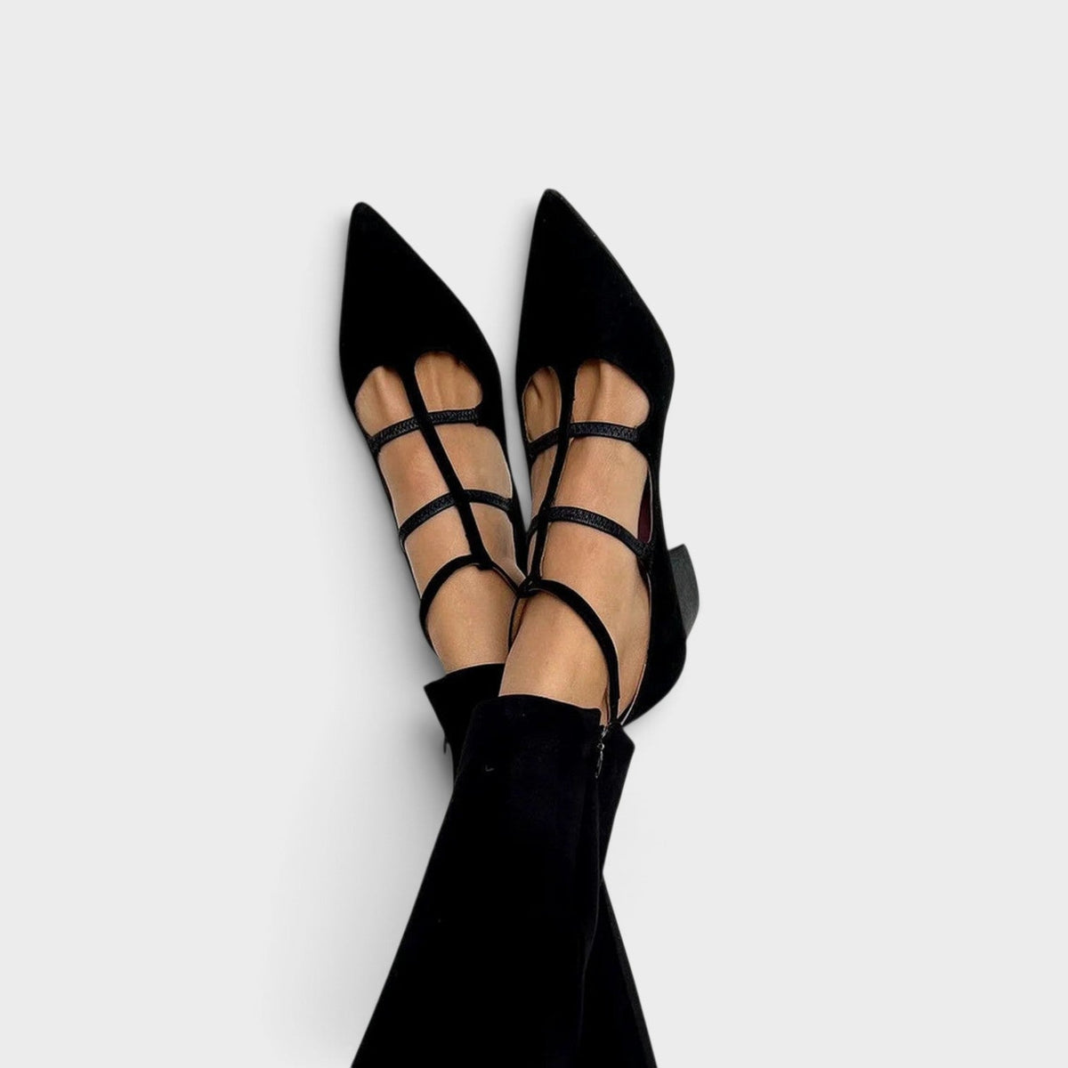 Getragene schwarze Pumps mit spitzer Kappe, mehreren schmalen Riemchen und Knöchelriemen, Beine gekreuzt vor hellem Hintergrund, dazu schwarze Hose. 1