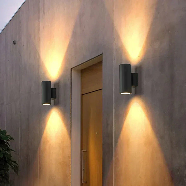 Zwei dunkle Wandleuchten beidseitig einer Holztür an einer Betonfassade, die Lichtkegel nach oben und unten erzeugen. 6