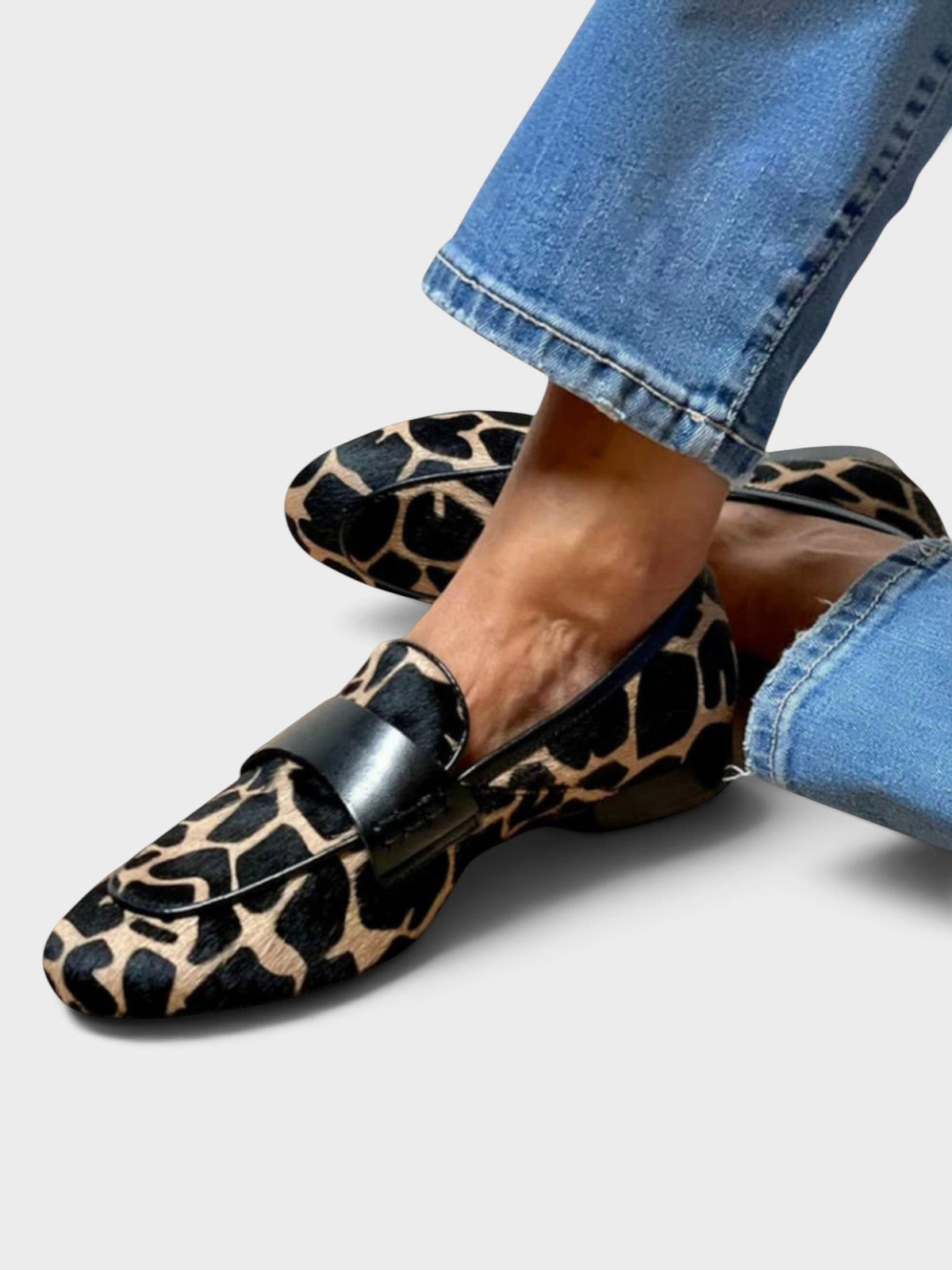 Flache Slip-on-Loafer mit schwarz-beigem Fellmuster und schwarzem Lederband, getragen zu Jeans, ein Fuß über den anderen gelegt. 1