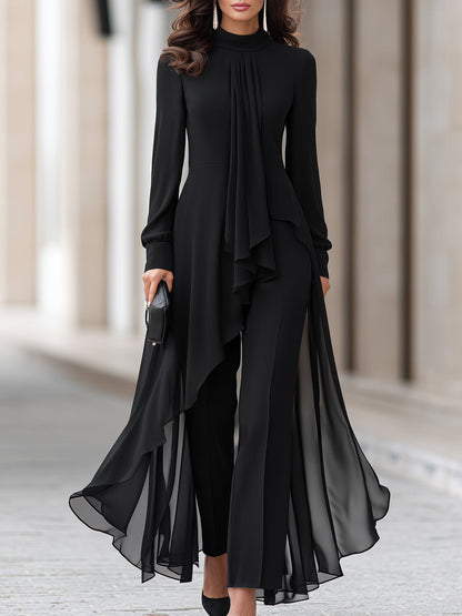Frau in schwarzer Chiffon-Tunika mit Stehkragen und weiter Hose, Halbtotale von der Taille abwärts, schwarze Clutch in der Hand. 1