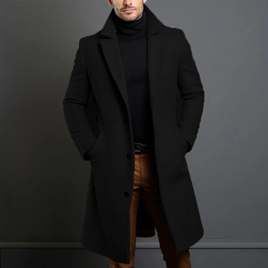 Model trägt den langen einreihigen Mantel in Schwarz mit Reverskragen zu schwarzem Rollkragenpullover und brauner Hose, Hände in den Taschen. 1
