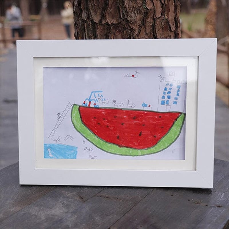 Weißer Bilderrahmen mit Passepartout auf einem Holztisch im Freien, darin eine Kinderzeichnung einer riesigen Wassermelone mit Gebäude und Figuren. 21