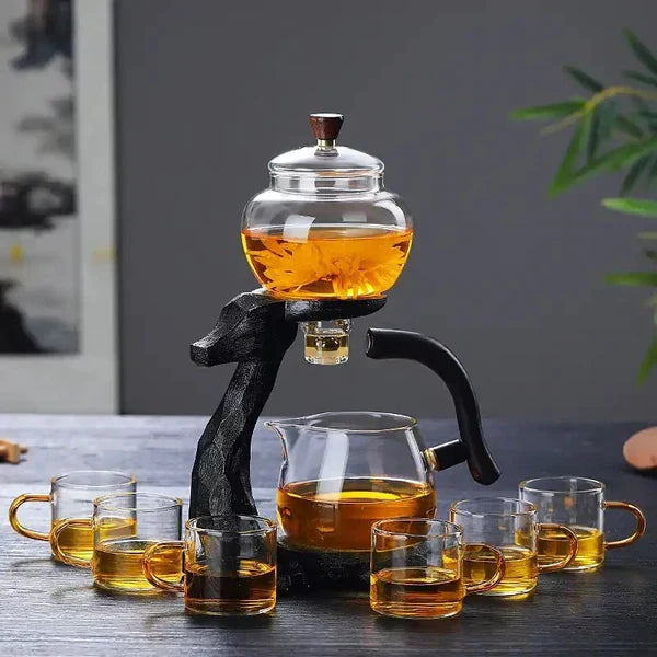 Teeservice mit Glas-Teekanne, schwarzem Ständer und sechs Glastassen auf Holztisch.