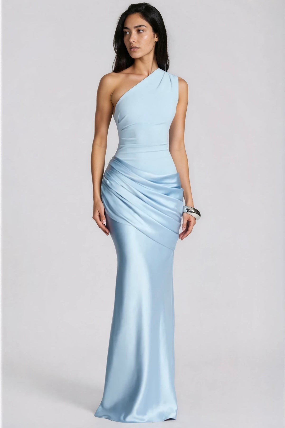 Model in hellblauem Maxikleid mit einer Schulter und gerafftem Satinrock, leicht zur Seite blickend, silberner Armreif, Ganzkörperansicht. 1