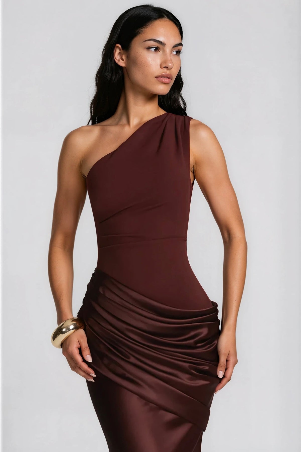 Nahaufnahme des Oberteils im dunkelbraunen Kleid mit Einschulter-Schnitt, Taillennaht und glänzend drapiertem Satin, goldener Armreif sichtbar. 9