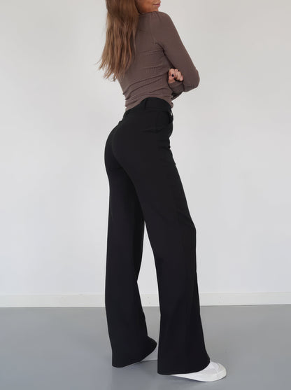 Frau in schwarzer Schlaghose und braunem Oberteil, seitlich, weiße Sneaker, minimalistischer Stil.