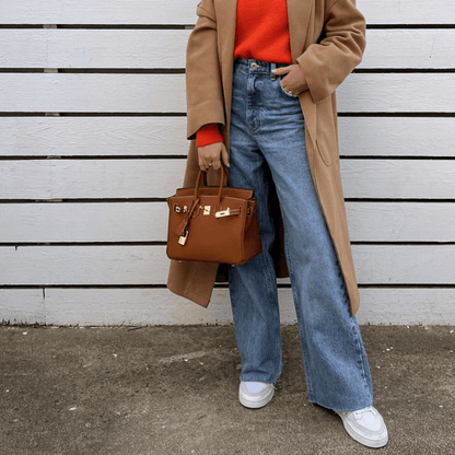 Frau in beigem Mantel, rotem Pullover, blauer Jeans, weißem Sneaker, brauner Handtasche.