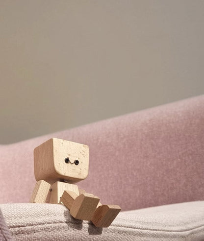 Kleine Holzfigur mit lächelndem Gesicht sitzt auf der Kante eines rosa Polstersofas vor einer grauen Wand. 4