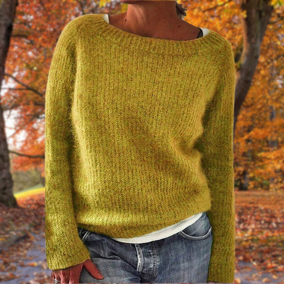 Senfgelber Strickpullover mit Rundhalsausschnitt, getragen über einem weißen Shirt und Jeans vor herbstlichem Laubwald. 1