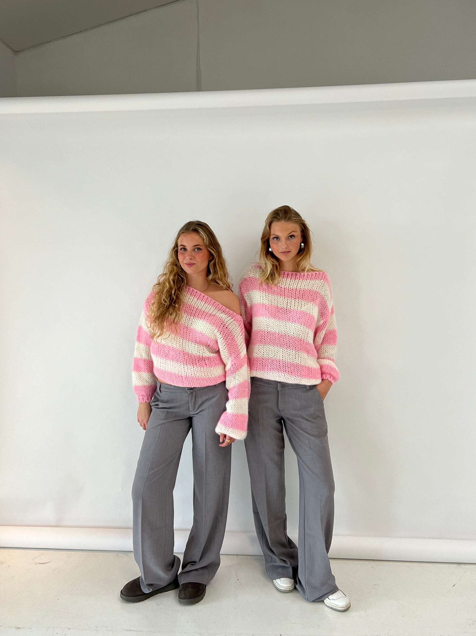 Zwei Frauen in gestreiften rosa Pullovern und grauen Hosen vor weißem Hintergrund.