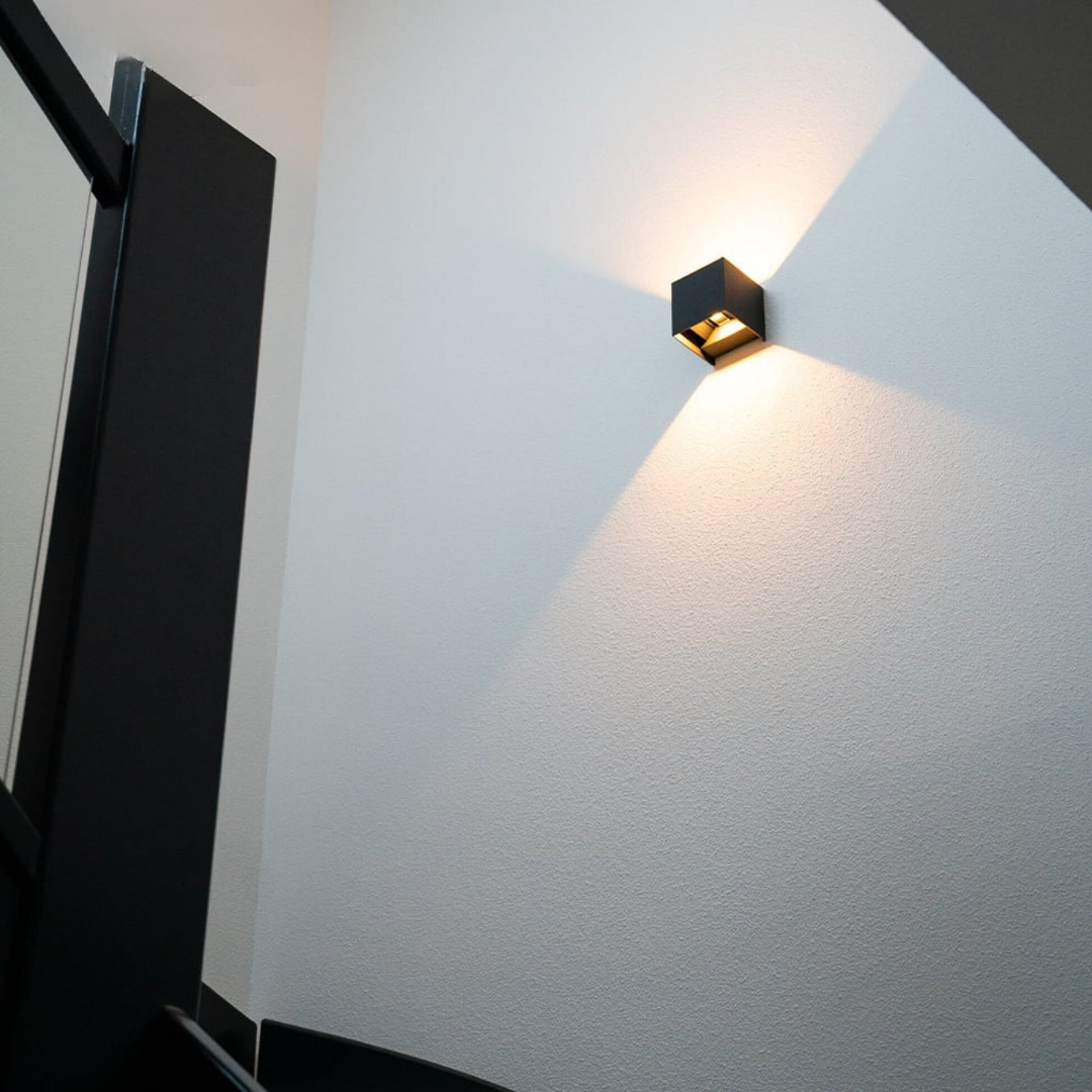 Schwarze Würfelleuchte an einer hellen Treppenhauswand, von unten fotografiert, mit Lichtkegeln nach oben und unten. 2